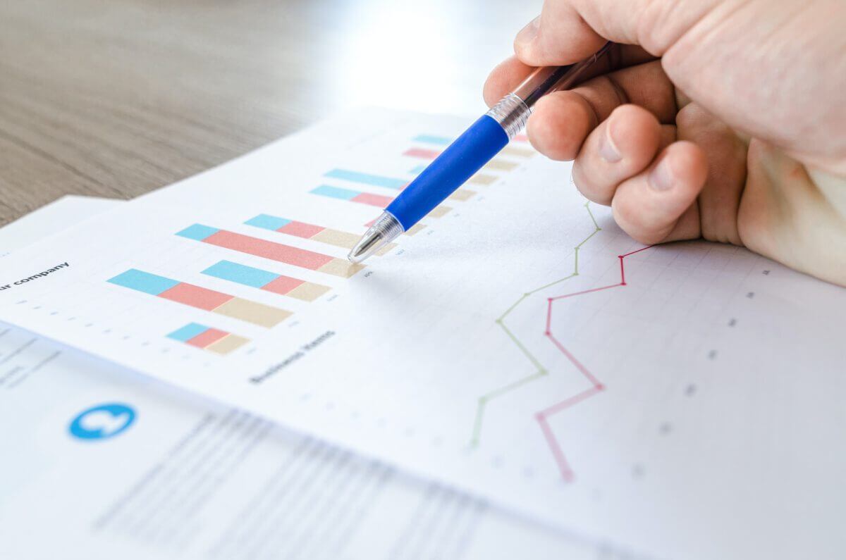 Closeup of a hand holding pen, touching a stack of papers with graphs and data visuals